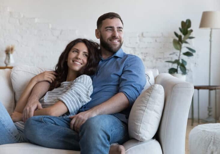 Cheerful dreamy young dating couple in love resting on sofa, relaxing at home, enjoying leisure, hugging, looking away, planning family future, marriage, mortgage, talking, laughing